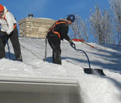 Professional technicians performing roof snow removal with safety harnesses and shovels in Des Moines, IA